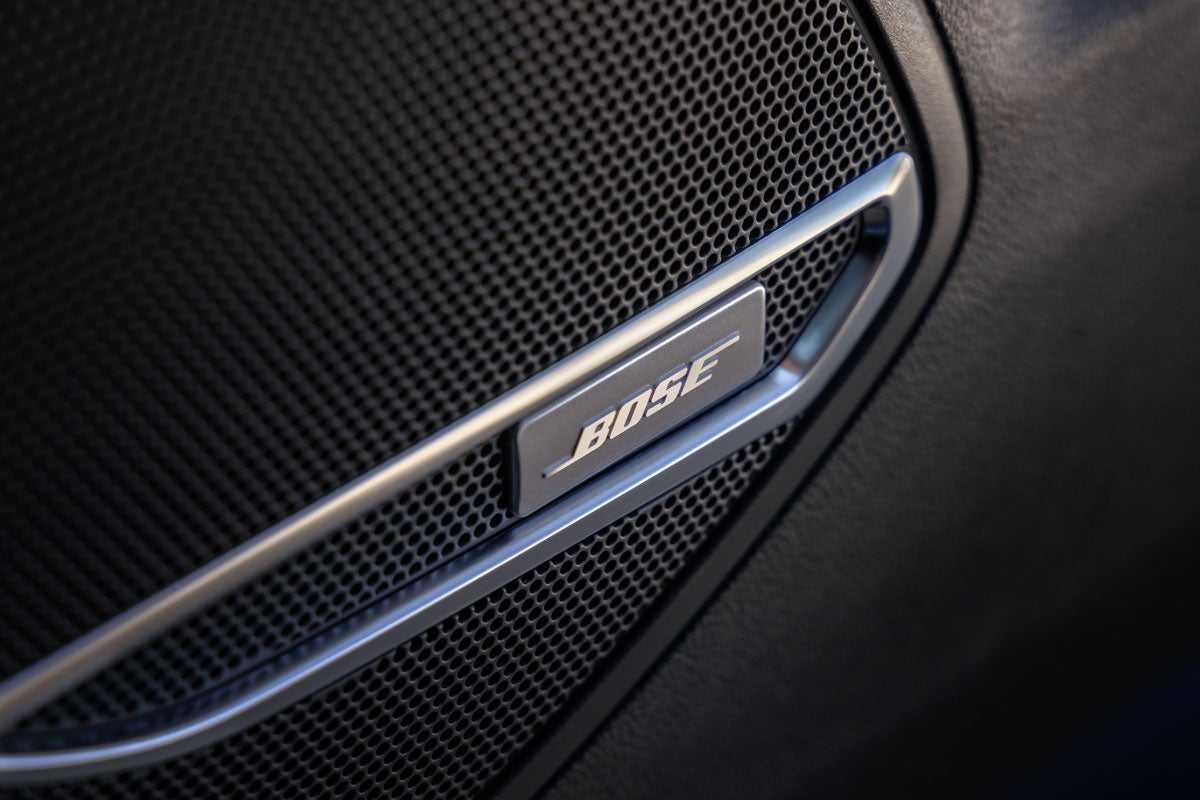 Close-up of a silver Bose logo on a car speaker grill, set against a dark, textured surface.