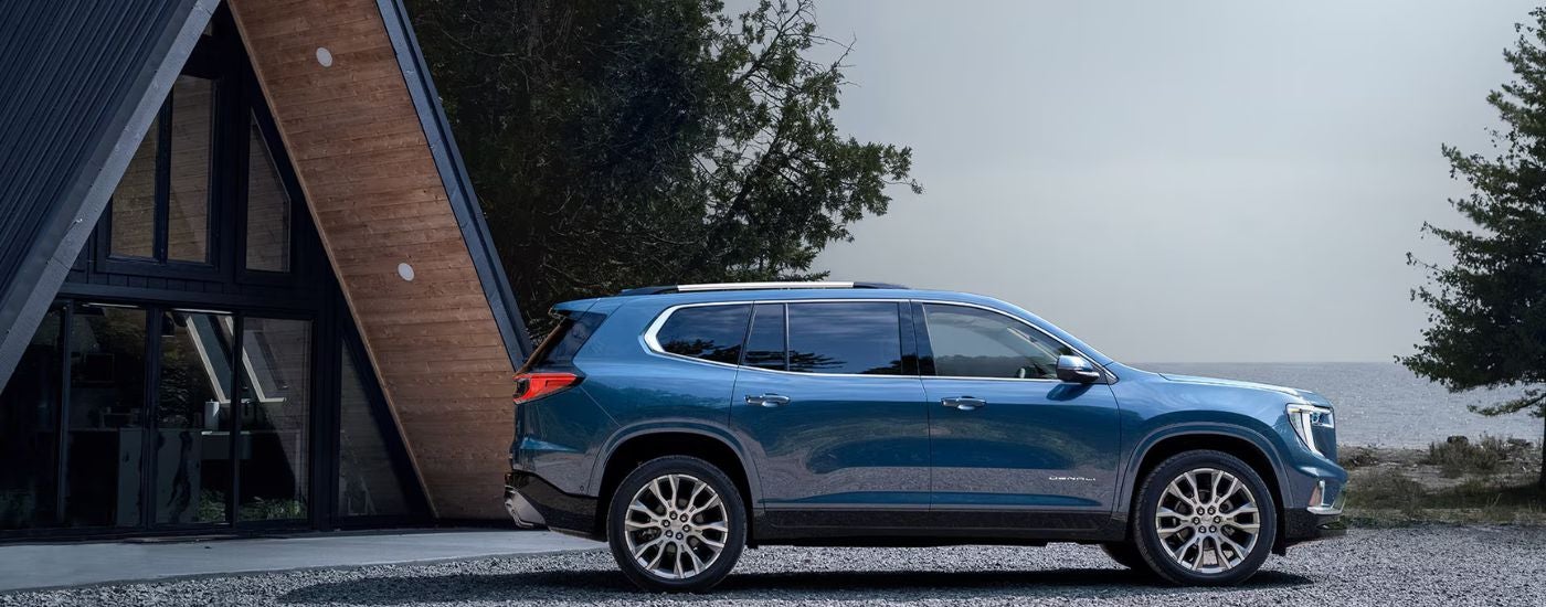 Blue 2026 GMC Acadia Denali parked near a lakeside cabin.