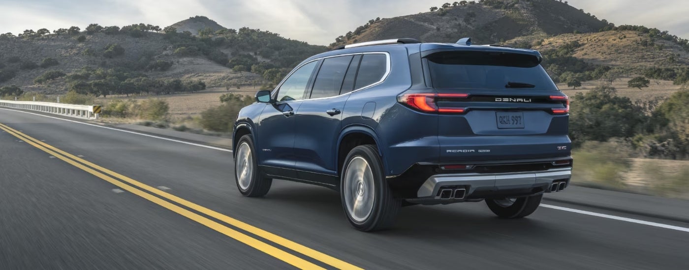 Rear view of a blue 2025 GMC Acadia Denali driving on a highway